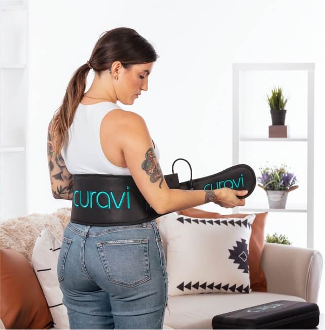 A young woman with tattoos using a laser light Curavi Belt for pain relief on her living room. 